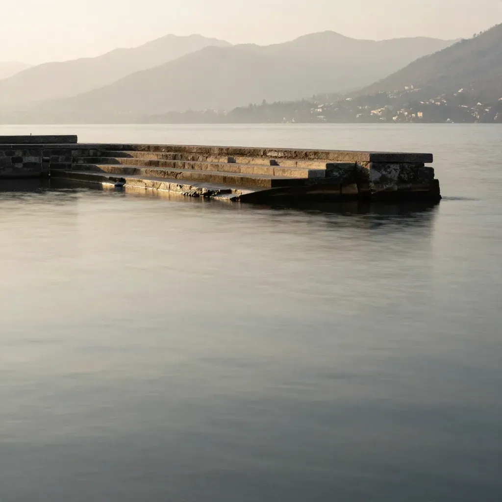 Il Lago di Como con riva di pietra e montagne lontane nella foschia calda.